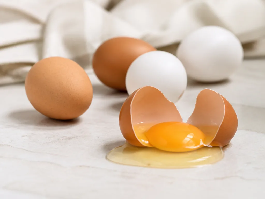 Färska ägg och ett uppknäckt ägg med synlig vita och gula på en ljus köksbänk
